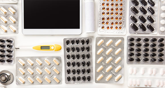 variety of medication tablets and capsules arranged around a digital thermometer and tablet device suitable for health monitoring and medication management